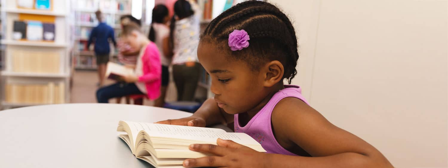 young girl reading a book sitting at a table in a classroom.