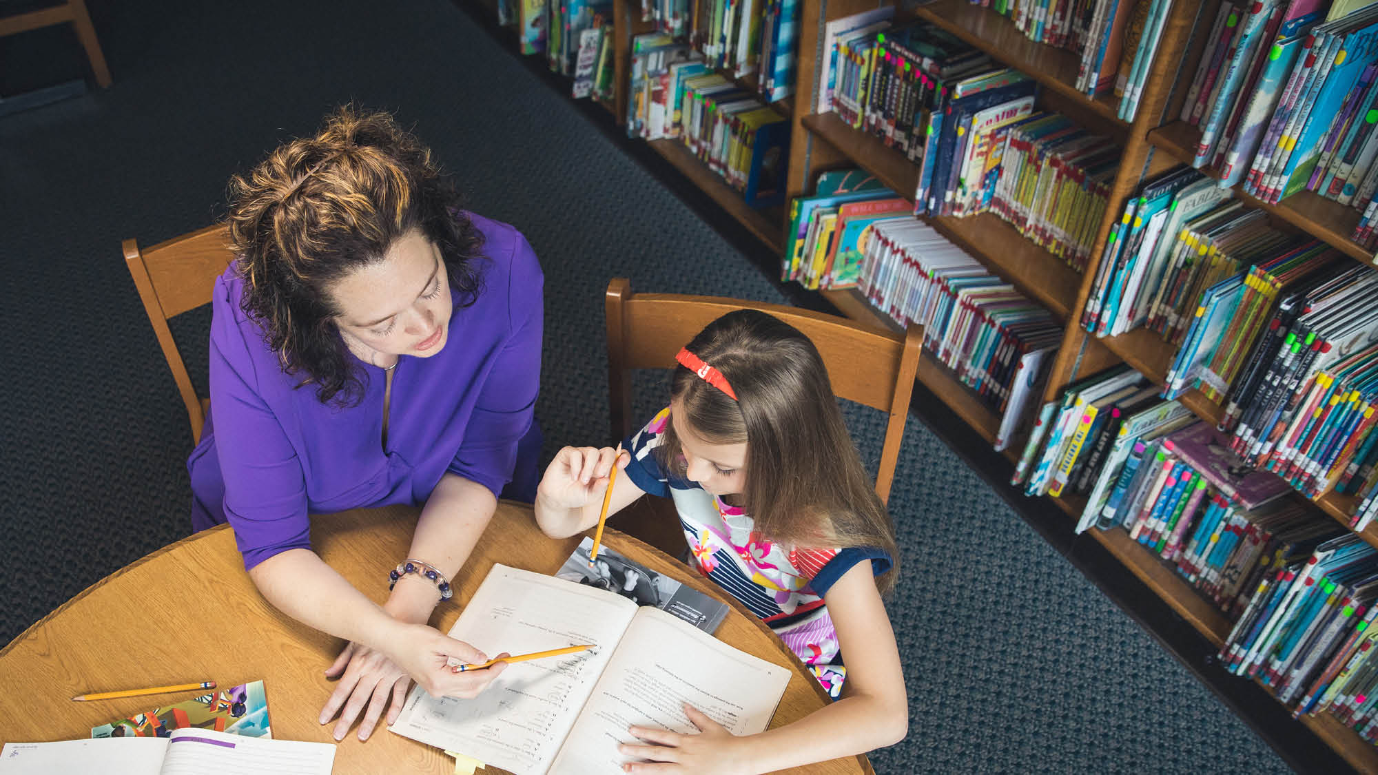 A teacher from Regent University's Career Switcher program sitting at a library table with a young student reviewing a workbook.